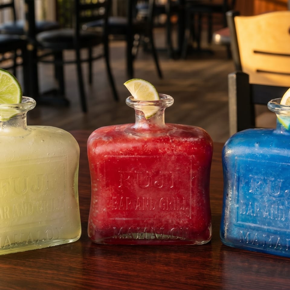 Three colorful frozen drinks in square glass bottles with citrus garnishes on a wooden table.