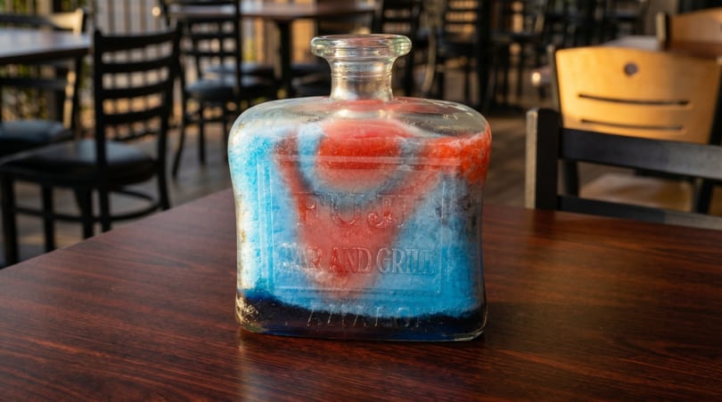 A glass bottle with red and blue slushy contents, labeled "FUJI BAR AND GRILL," on a wooden table.